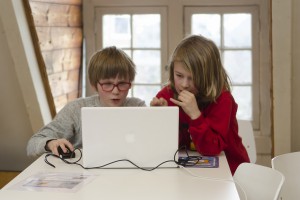 Two children sit at a white table, collaborating on a laptop as part of visual therapy. One uses the mouse while the other points at the screen, both focused on their task in a bright room with large windows.